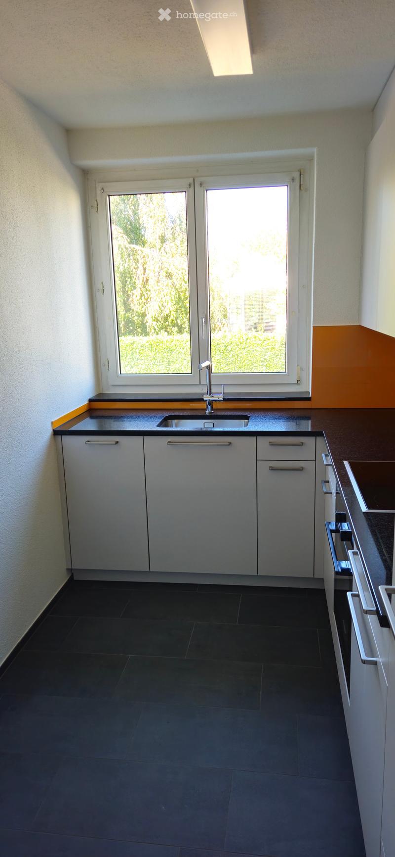 Modern kitchen with grey cabinets, black countertop, and a stainless steel sink under a large window.