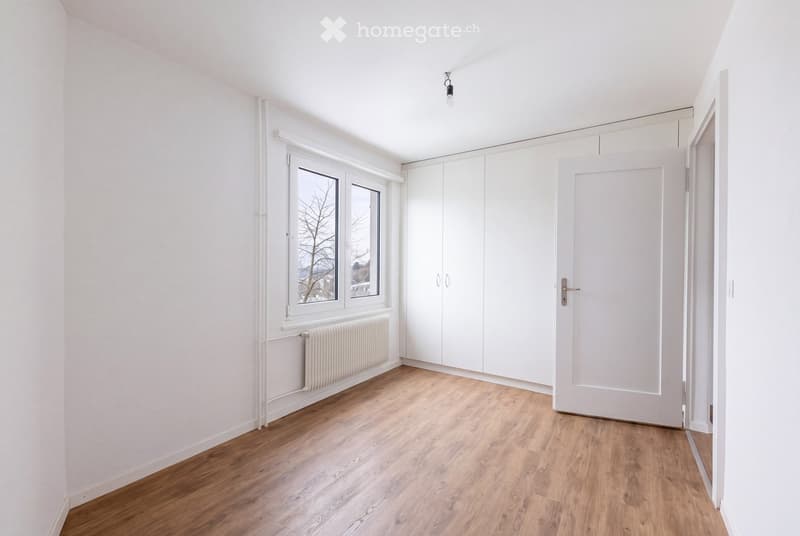 Empty room with white walls, wooden flooring, large windows, built-in white closet, and a radiator under the windows.