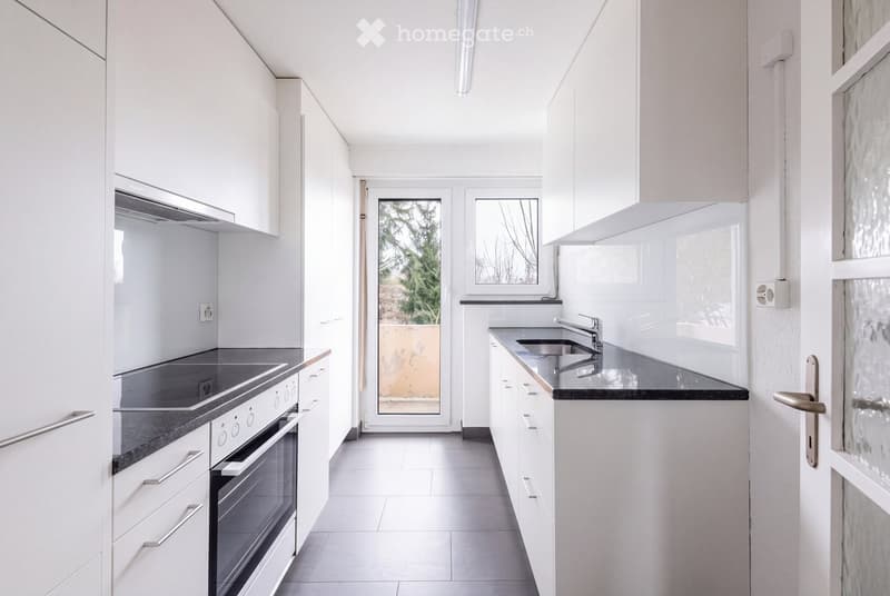 Modern kitchen with white cabinets, black countertops, built-in oven, and a sink with a modern faucet. The space is bright with large windows and a door leading outside.