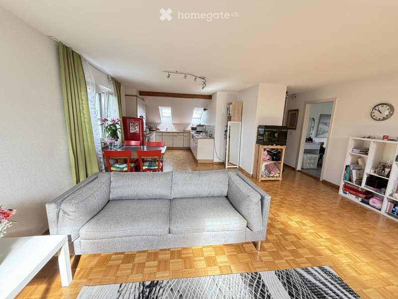 A modern living room with a grey sectional sofa, a white coffee table, a dining area with red chairs, a kitchen area with white cabinets and a red refrigerator, and a fish tank on a wooden shelf.