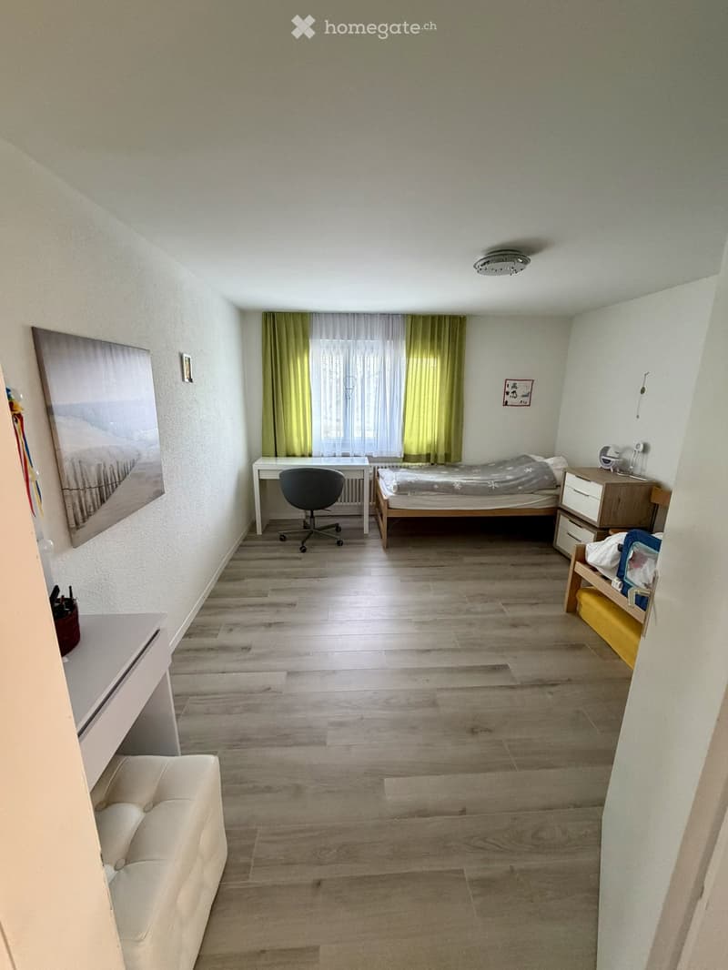 Cozy bedroom with wooden flooring, a bed, a desk, a chair, a nightstand, and a sofa. The room features large windows with curtains and a picture on the wall.