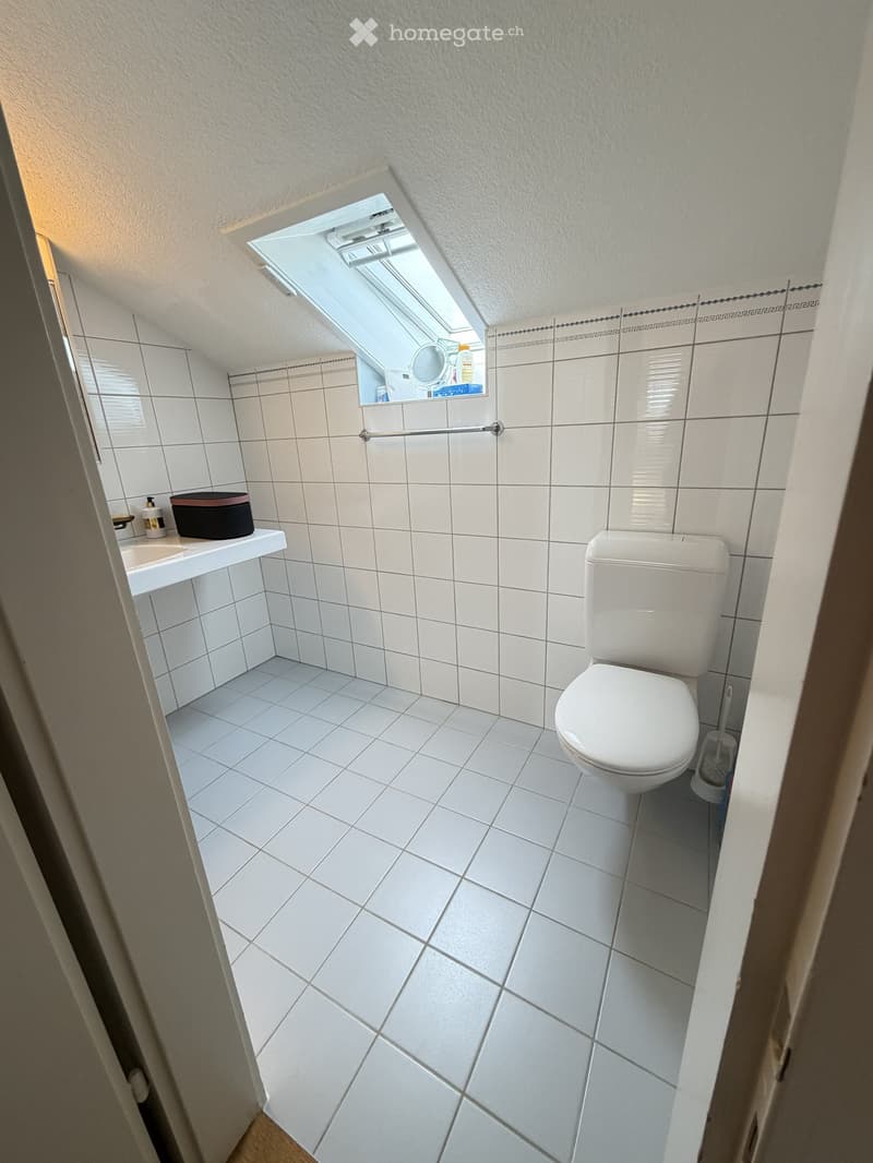 Small bathroom with white tiled walls and floor, a toilet, a sink with a mirror above it, and a small window at the top.