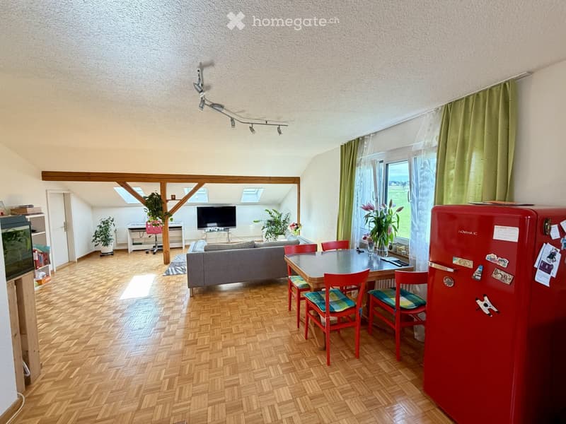 Bright, open living and dining area with wooden flooring, a grey sofa, a dining table with red chairs, a red refrigerator, and a television in the background. Large windows with green curtains let in natural light.