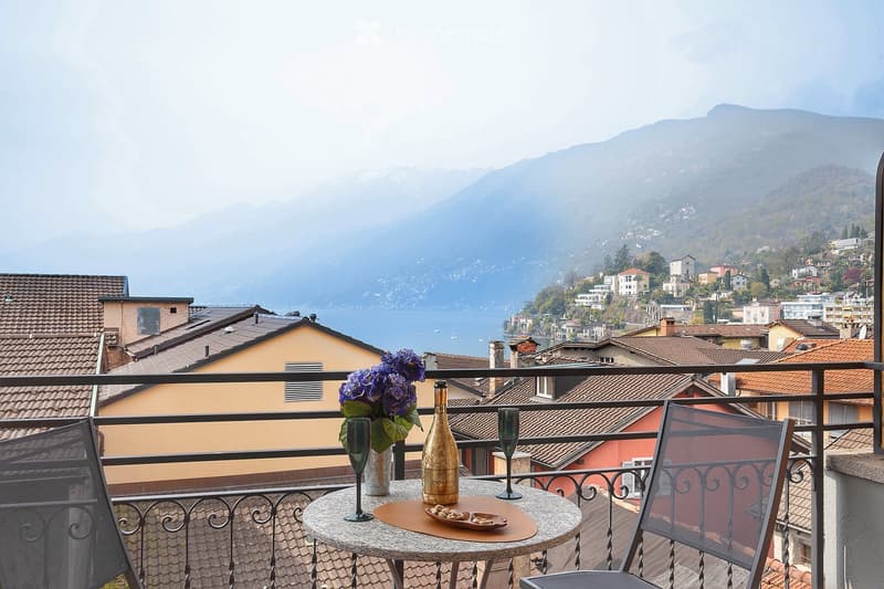 Balcony with a scenic view of the mountainous landscape, featuring a table with a vase of flowers, a bottle of wine, and two wine glasses. The balcony overlooks a town with colorful buildings and rooftops in the distance.