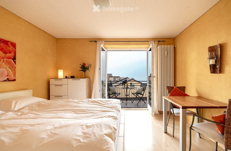 Spacious bedroom with a balcony, wooden furniture including a dresser and a table, and a large window offering a scenic view.