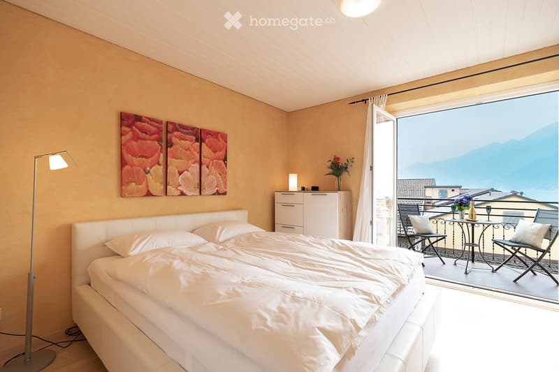 Spacious bedroom with a large balcony, beige walls, a white bed with bedding, a dresser, and artwork on the walls