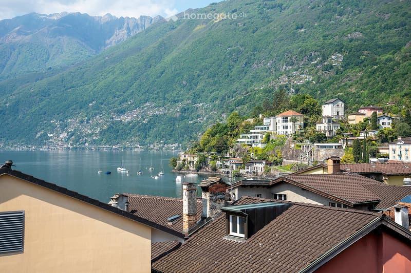 The image shows a scenic view of a lakeside town nestled among lush, green mountains. The town features a mix of residential and commercial buildings with red-tiled roofs, and there are several boats and yachts anchored in the calm waters of the lake. The