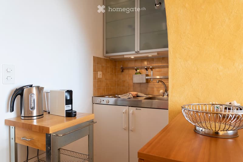 Small kitchen with tile backsplash, stainless steel appliances including a dishwasher, and a wooden countertop with a kettle and coffee maker on it.