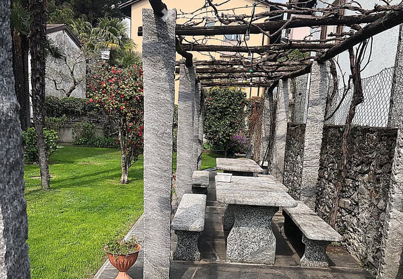 Lush garden with green grass, flowering plants, stone pathway, and a stone fireplace structure with a pergola overhead