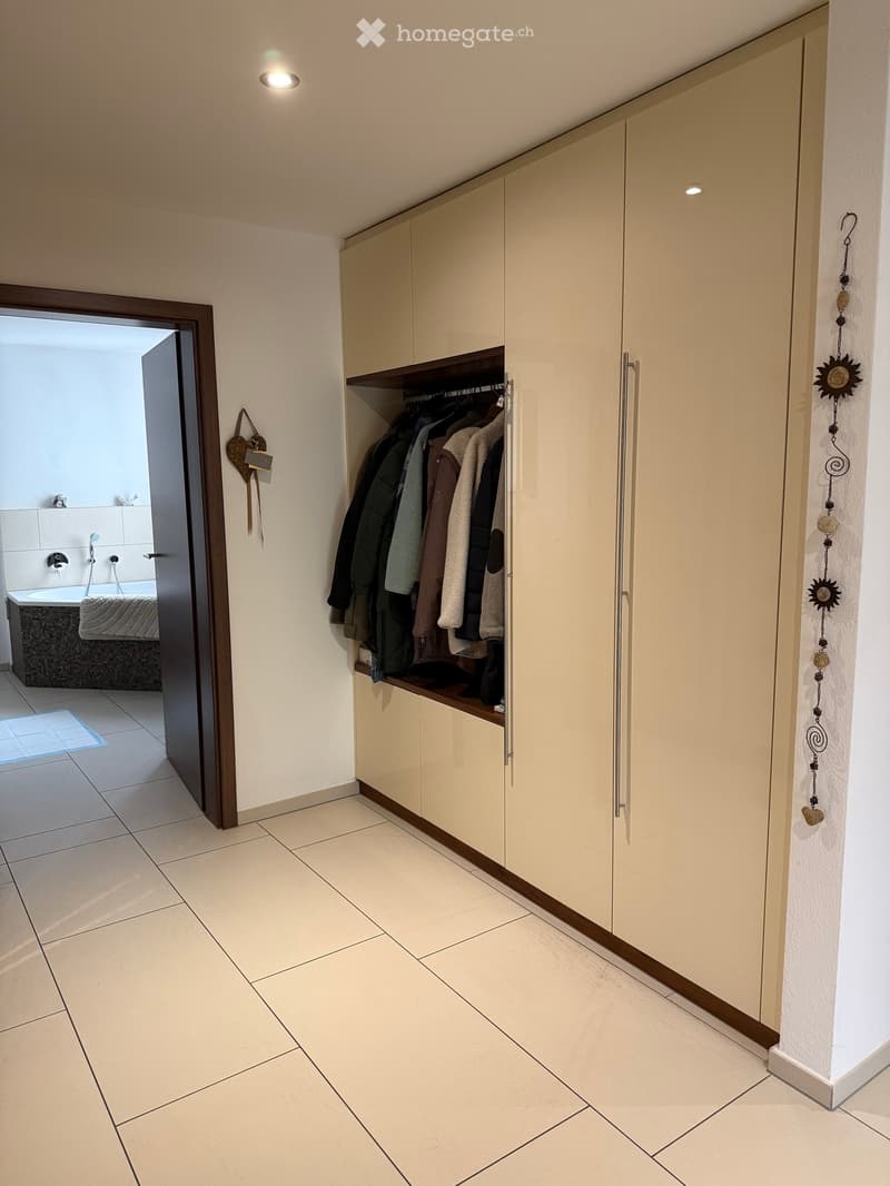 Hallway with tiled floor, built-in wardrobe with hanging clothes, decorative wall hanging, and an open doorway leading to a bathroom with a bathtub and shower.
