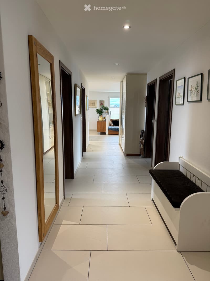 Clean, well-lit hallway with tiled flooring, white walls, and several doors leading to different rooms. Features include a bench with a cushion, framed artwork, and a mirror on the left side.