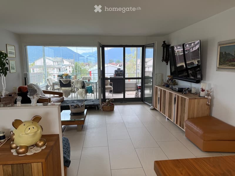 Bright living room with large sliding glass doors leading to a balcony, offering a scenic mountain view. The room features a sofa, coffee table, ottoman, chairs, a television, cabinets, and decorative items including a plant and a ceramic pig figurine.
