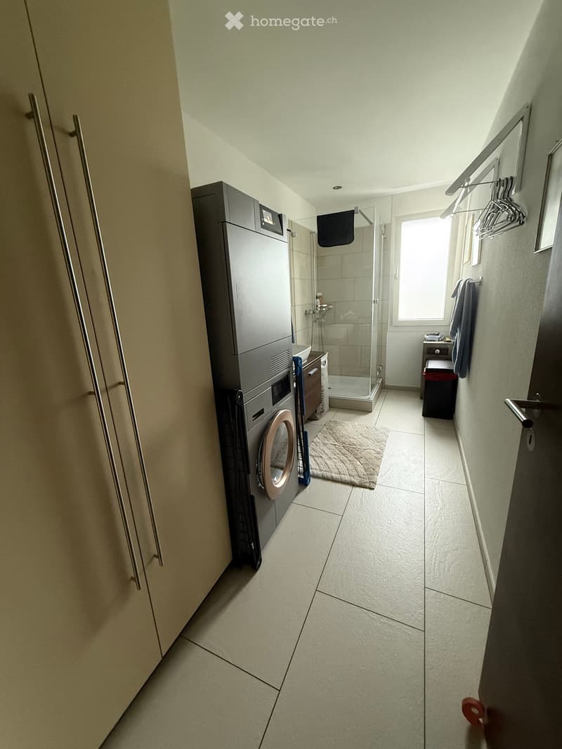A narrow hallway with tiled flooring, a washing machine, a shower stall, a small table with a chair, and a coat rack with towels.
