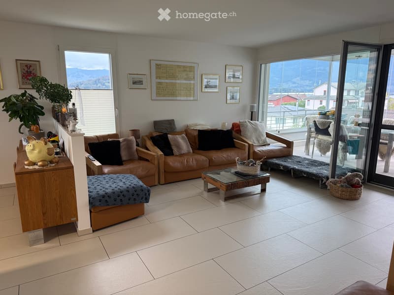 Bright living room with large windows and a view, featuring a brown leather sofa, glass coffee table, ottoman, and decorative plant on a side table.
