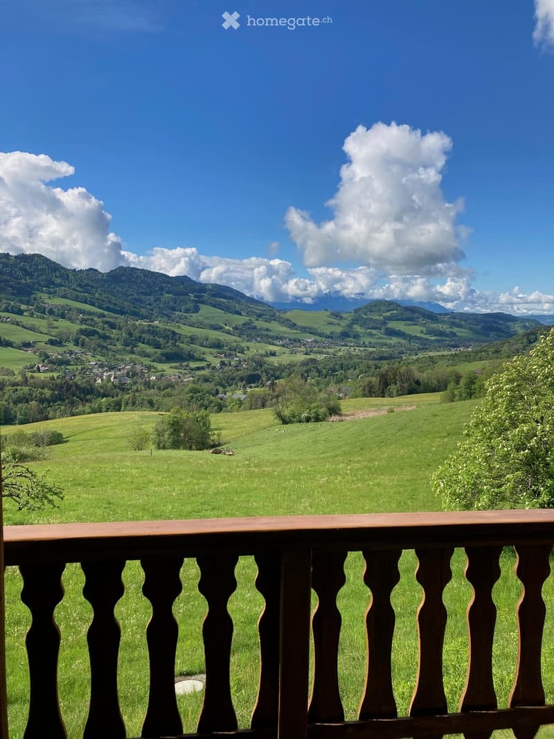 Balcony overlooking a lush green valley with rolling hills, scattered trees, and distant mountains under a partly cloudy sky.