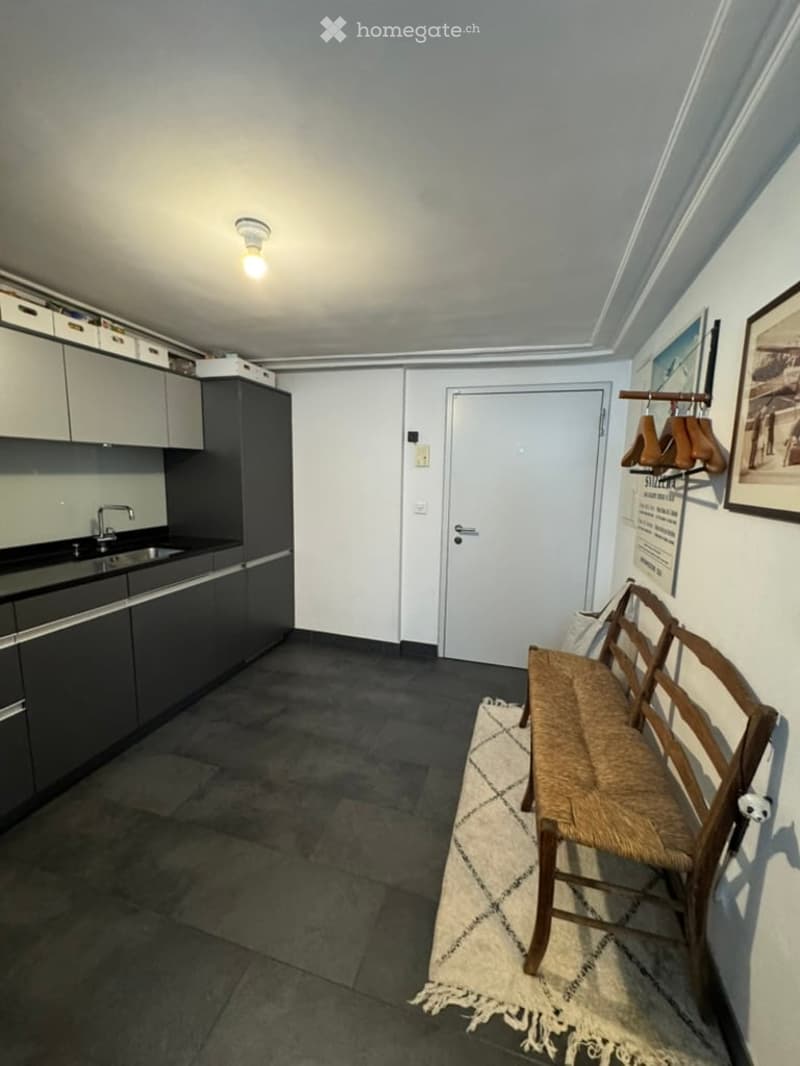 A small, modern kitchen area with grey cabinets, a sink, and a wooden bench with a woven seat. A coat rack with hangers and a picture frame are visible on the wall.