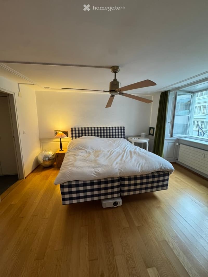 Cozy bedroom with wooden flooring, a large bed with checkered headboard, a nightstand with a lamp, a small round table, and a decorative ball on the floor.