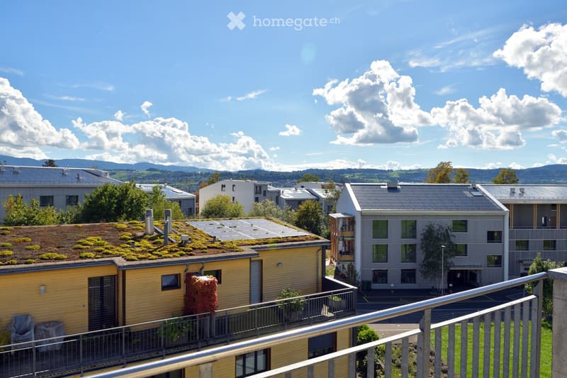 View of a residential complex with modern buildings, green roofs, and surrounding greenery under a clear blue sky.