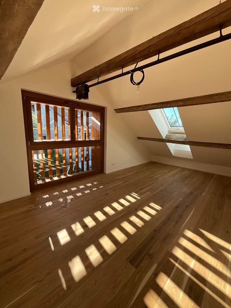 Bright, empty attic space with wooden flooring, large skylight, and a large window with wooden frame. Exposed wooden beams on the ceiling.