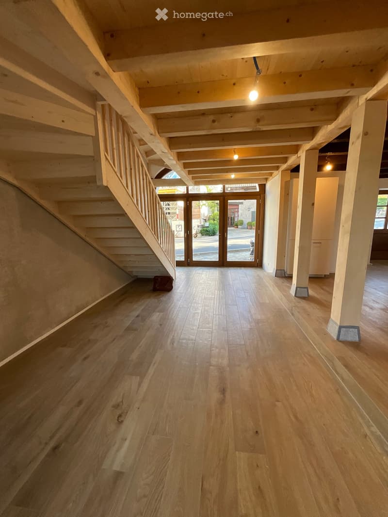 Empty hallway with wooden flooring, exposed wooden beams on the ceiling, and a staircase leading upwards. Natural light streams in from the glass doors at the far end.
