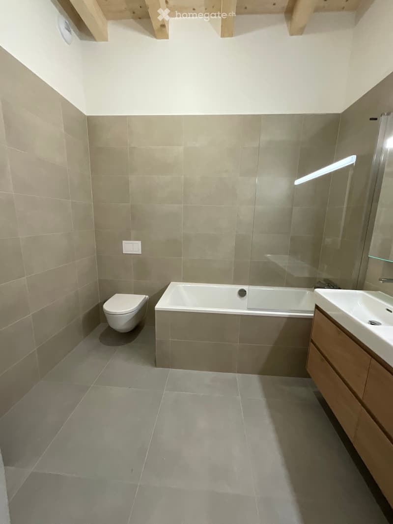 Modern bathroom with a bathtub, toilet, sink, and wooden cabinet. Light grey tiles on walls and floor, sleek design.