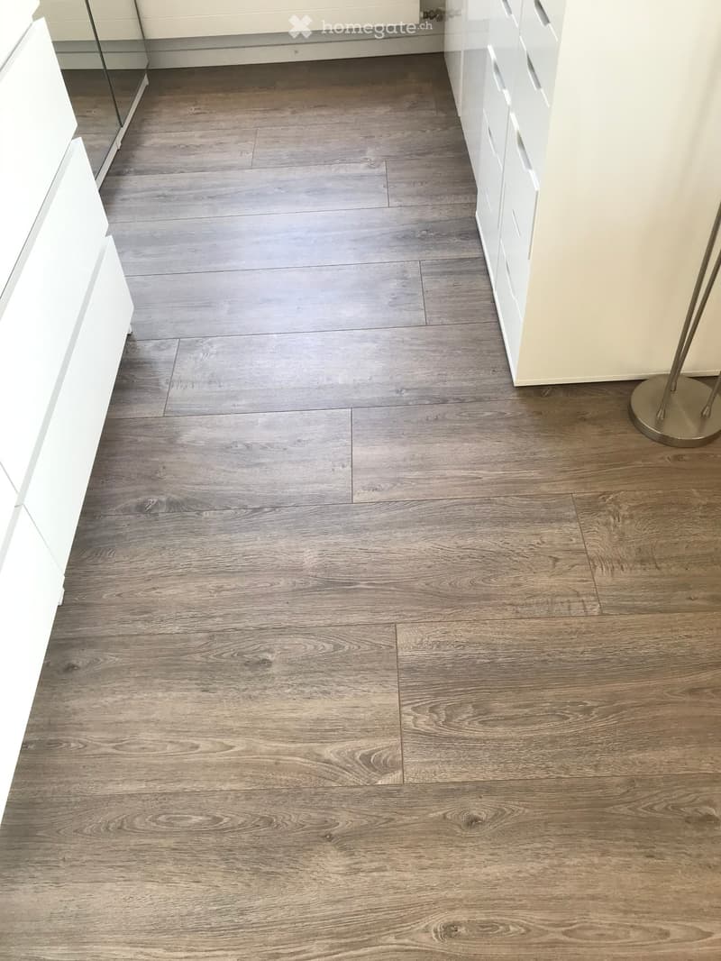 Empty room with grey wooden flooring, white walls, and a small section of white cabinetry visible on the left side.