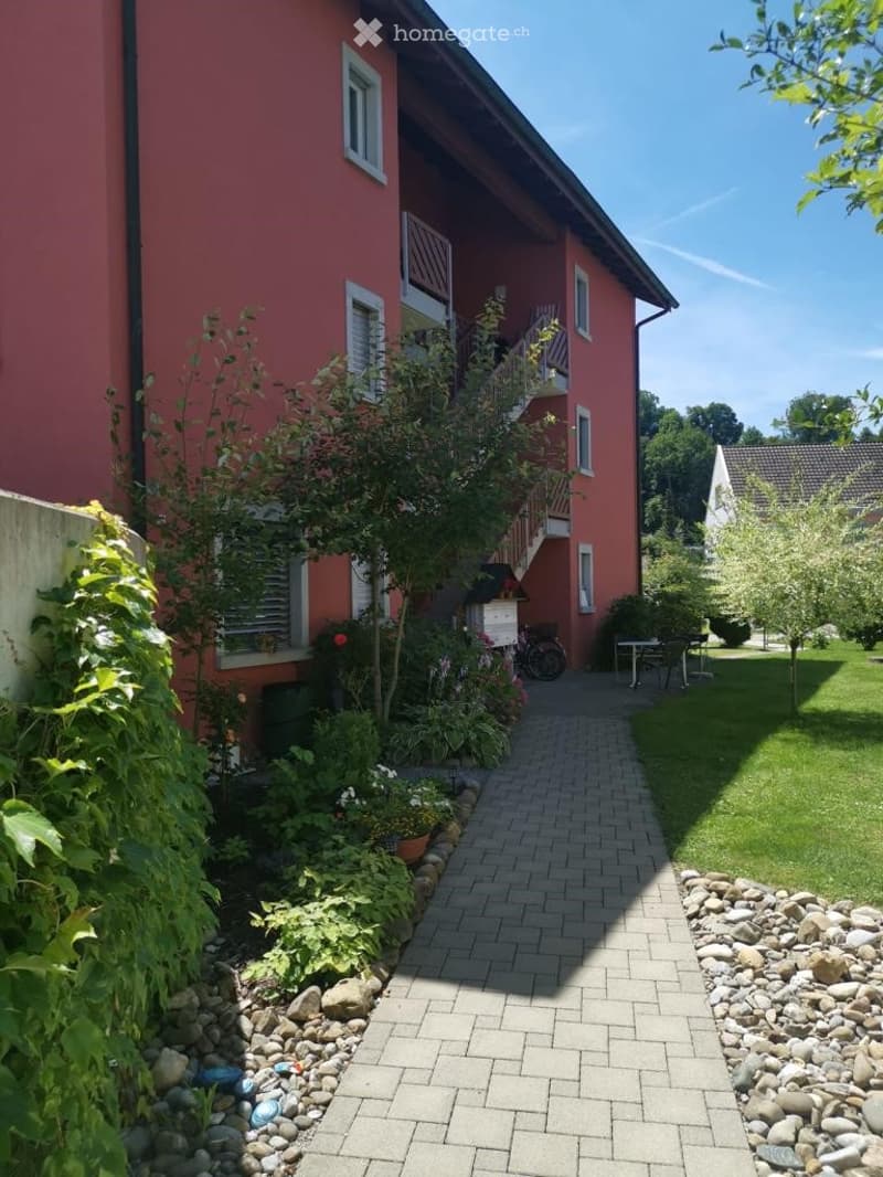 Red brick two-story building with white-framed windows, a staircase leading to a balcony, and a well-maintained garden with a brick pathway, small table, and chairs.