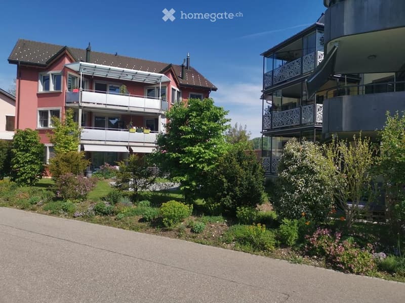 Two-story residential building with balconies, surrounded by greenery and a well-maintained garden.