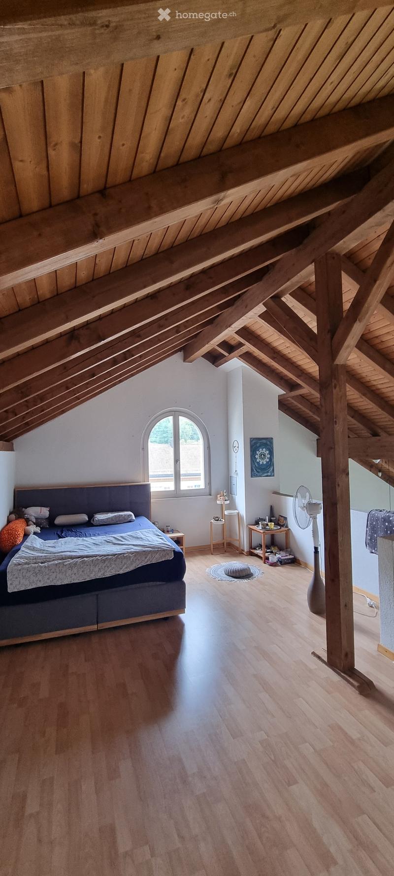 Cozy attic bedroom with wooden beams, hardwood floor, large window, bed with pillows and blankets, nightstand, fan, small table, and decorative items.