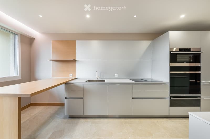 Modern kitchen with white cabinets, a central island, and built-in ovens. Features a sleek design with a light-colored countertop and a large window on the left side.