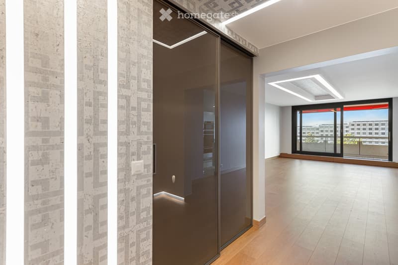 Empty room with wooden flooring, white walls, large glass sliding door leading to a balcony, and modern lighting fixtures.