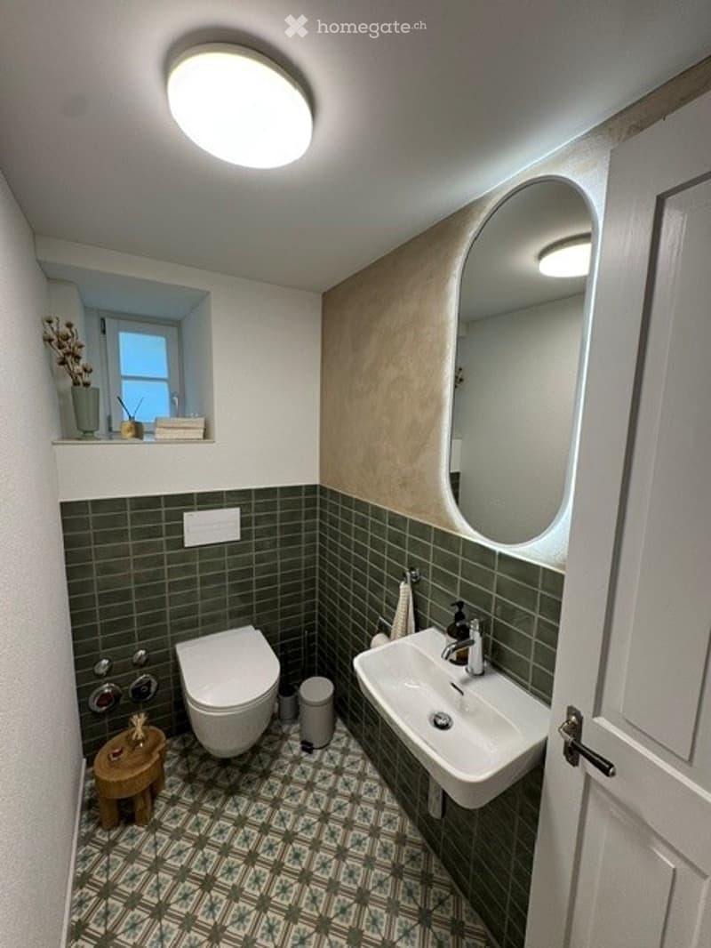 Modern bathroom with green tiles, white fixtures, a round mirror, a small wooden stool, and a vase with dried flowers on the windowsill.