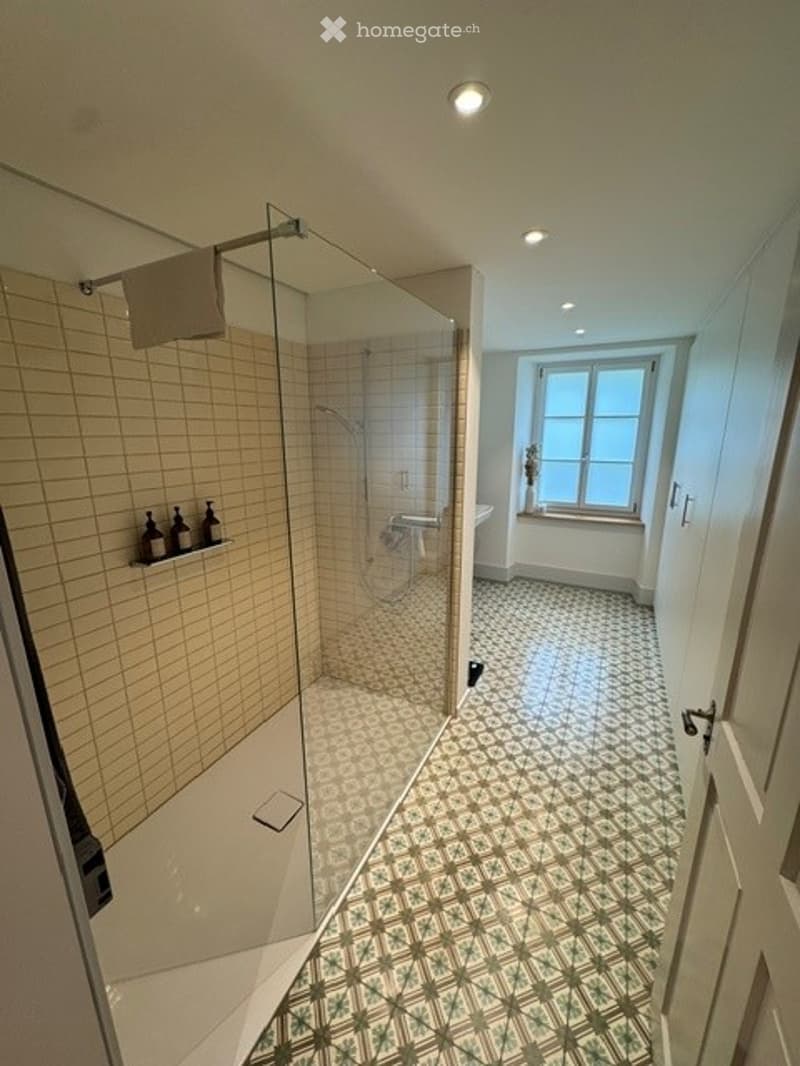 Modern bathroom with a glass-enclosed shower, tiled walls, and a bathtub near a window. The floor features a patterned tile design, and there are decorative bottles and a towel visible.