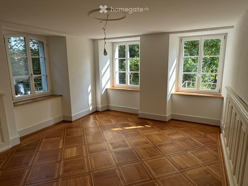 Empty room with wooden flooring, white walls, and three large windows allowing natural light to enter. The room features a simple, clean design with no furniture or decorations.