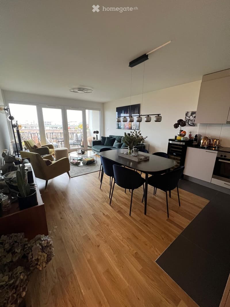 Modern living room and dining area with wooden flooring, large windows, sofa, coffee table, armchairs, ottoman, dining table with chairs, kitchen area with cabinets and oven, and decorative plants.