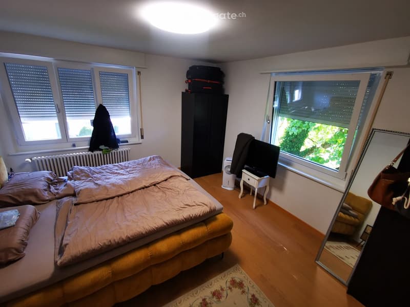Cozy bedroom with a large bed, wardrobe, small table with a fan, and a mirror. The room has wooden flooring and large windows with blinds.