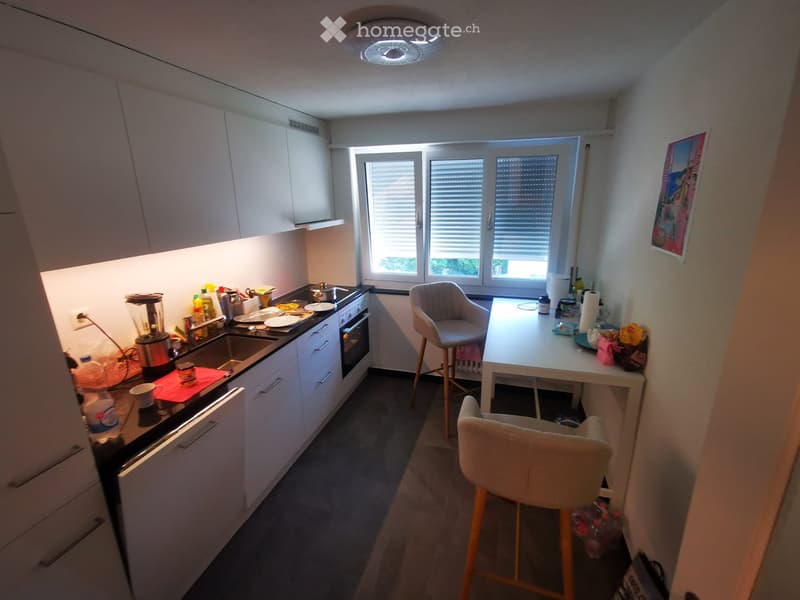 Modern kitchen with white cabinets, a sink, oven, and a small dining area with a table and two chairs. The counter has various kitchen items including a blender, cups, and plates.