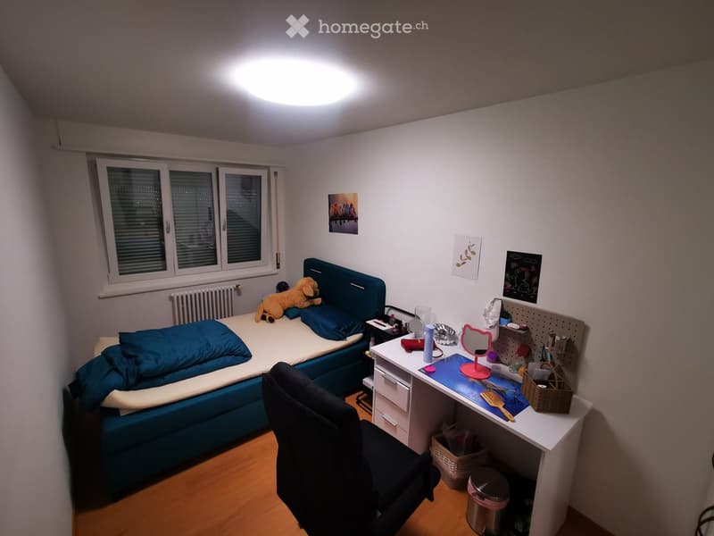 Cozy bedroom with a single bed, desk, chair, and various personal items. The room features a large window with blinds, a radiator, and several wall decorations.