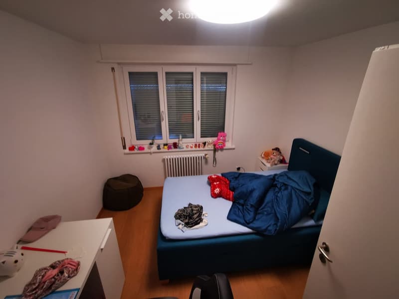 Cozy bedroom with a single bed, a small table, a round stool, and a window with blinds. The bed is unmade with a blue duvet and a red blanket. A small shelf above the radiator holds various items, including a teddy bear and other small objects.