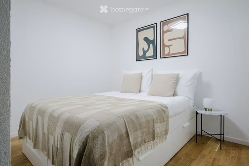 A modern bedroom with a neatly made bed, a nightstand with a lamp, and two picture frames on the wall.