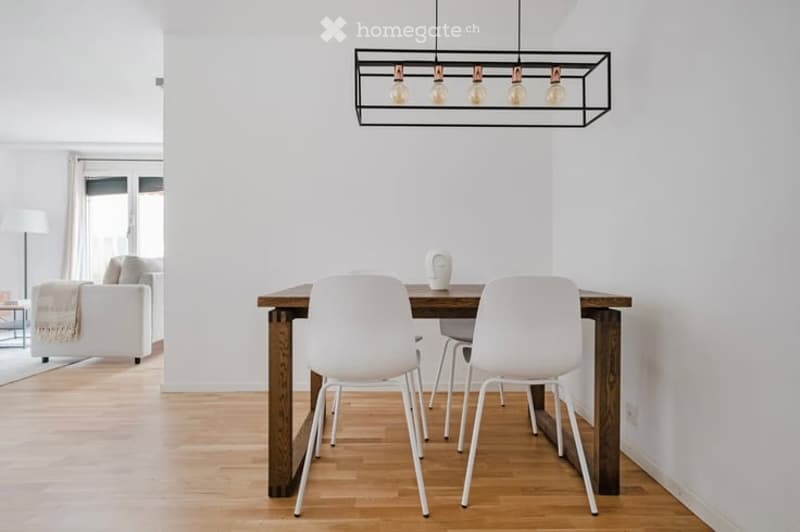 Bright, modern dining area with a wooden table, white chairs, and a decorative vase. Adjacent living area features a white sofa, coffee table, and floor lamp.