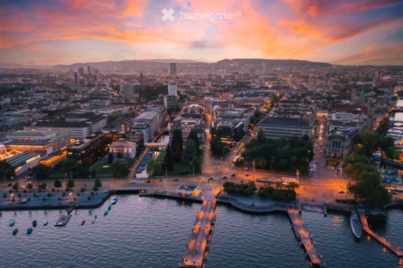 Aerial view of a cityscape at sunset, featuring a waterfront with docks, boats, and a mix of modern and historic buildings.