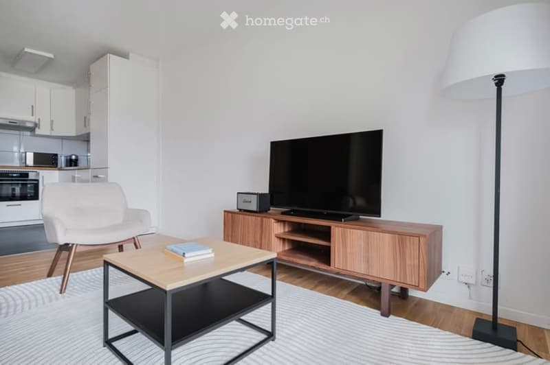 Modern living room with a white armchair, wooden coffee table, TV stand, floor lamp, and books on the table.