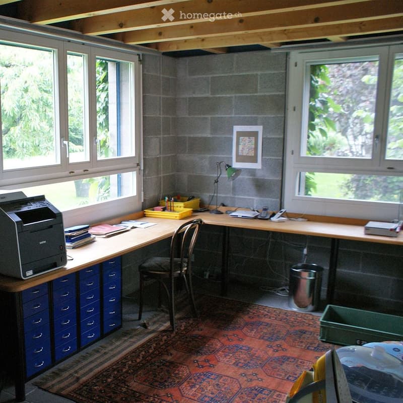 A well-lit room with exposed wooden beams, a large wooden desk, a chair, a printer, a lamp, a framed picture on the wall, and a colorful rug on the floor.