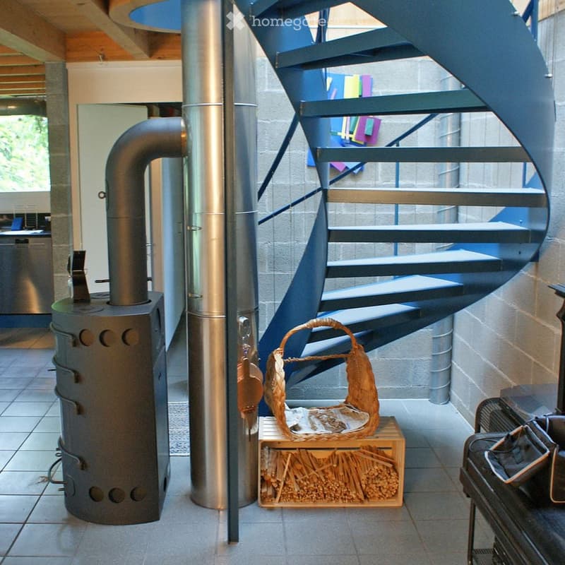 Modern hallway with a spiral staircase, wood-burning stove, and a basket of firewood on a wooden crate.