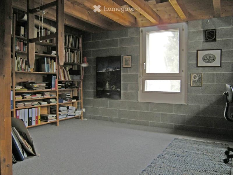 A cozy room with exposed wooden beams, a large bookshelf filled with books, a small table with a lamp, and a comfortable chair near a large window.