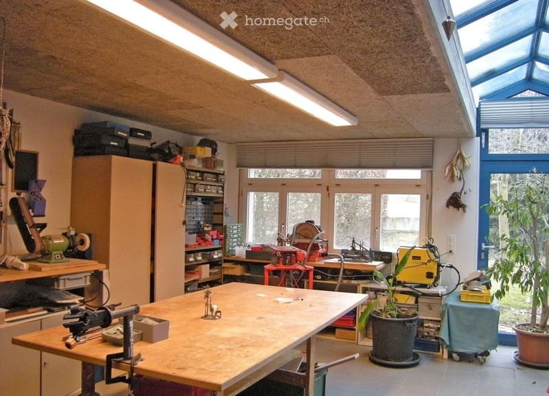 Well-lit workshop with large windows, a central workbench, various tools, cabinets, and potted plants.