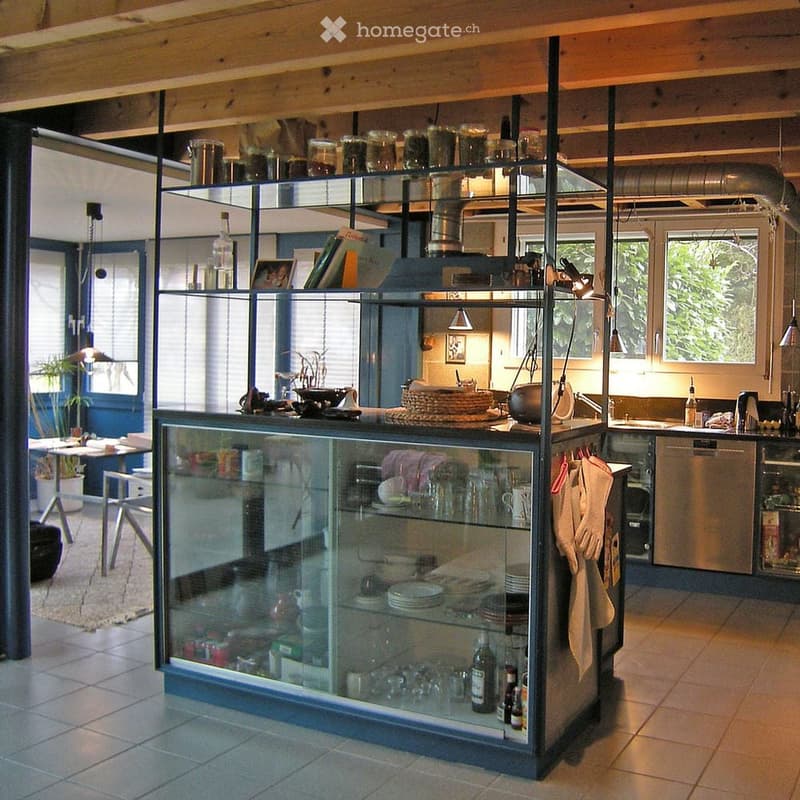 Modern kitchen with stainless steel appliances, glass display cabinet, and wooden beams on the ceiling. Includes a sink, dishwasher, oven, and various kitchen items on shelves and counters.
