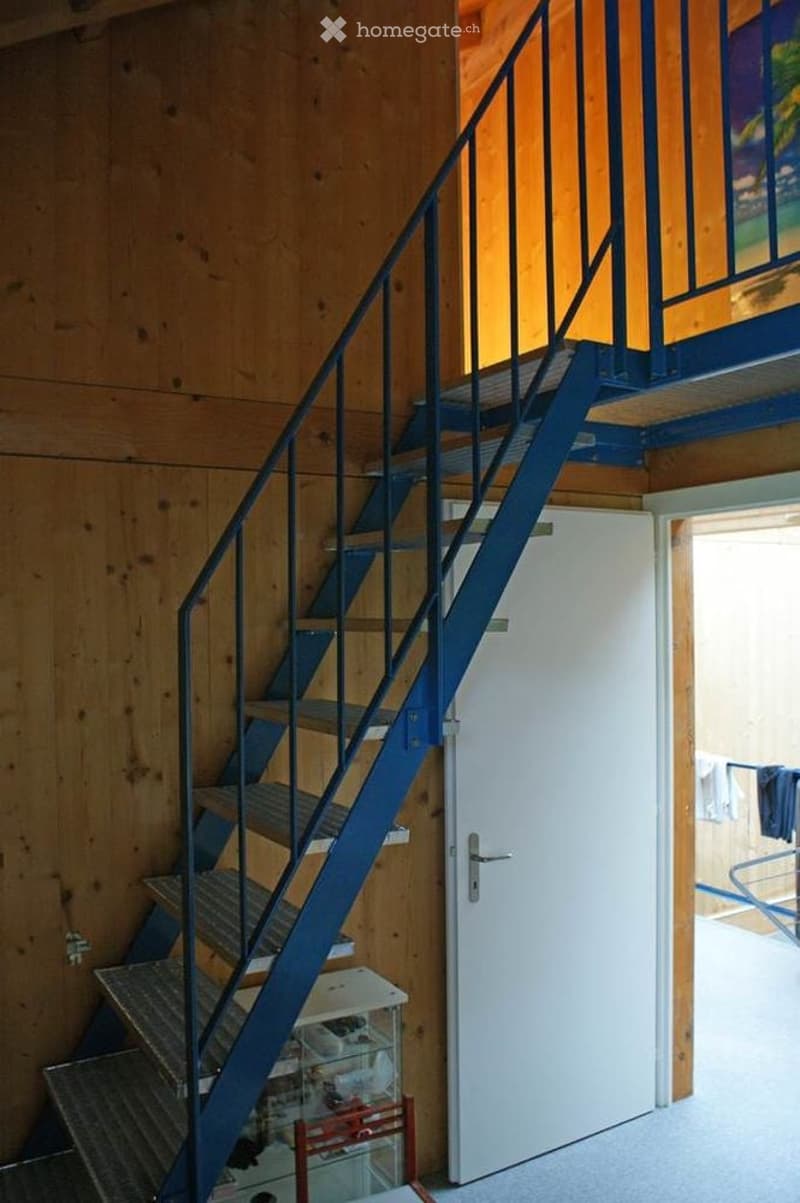 Wooden-walled hallway with a modern staircase featuring blue railings and steps, leading to an upper level. A white door is visible on the right side, and a small storage unit is placed near the base of the stairs.