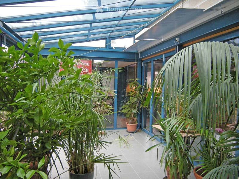 A covered outdoor area with a glass roof and blue frame, filled with various potted plants and greenery, including palms and leafy plants, with a tiled floor and glass doors leading to an indoor space.